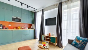 Uma sala de estar moderna com um sofá, uma mesinha com uma garrafa e copos, e uma área de cozinha com armários verde-água e um micro-ondas.