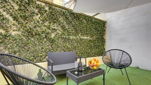 Área externa com assentos de vime, uma mesa com uma fruteira e uma garrafa, e uma parede coberta por plantas trepadeiras.