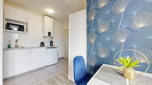 Cocina moderna con armarios blancos, microondas y cafetera. Hay una silla azul y una pequeña mesa con una planta.