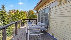 A wooden deck with a white outdoor seating area, a large window, and a view of trees and a house in the background. - Lake View Condo - Walk to Saugatuck (Douglas)
