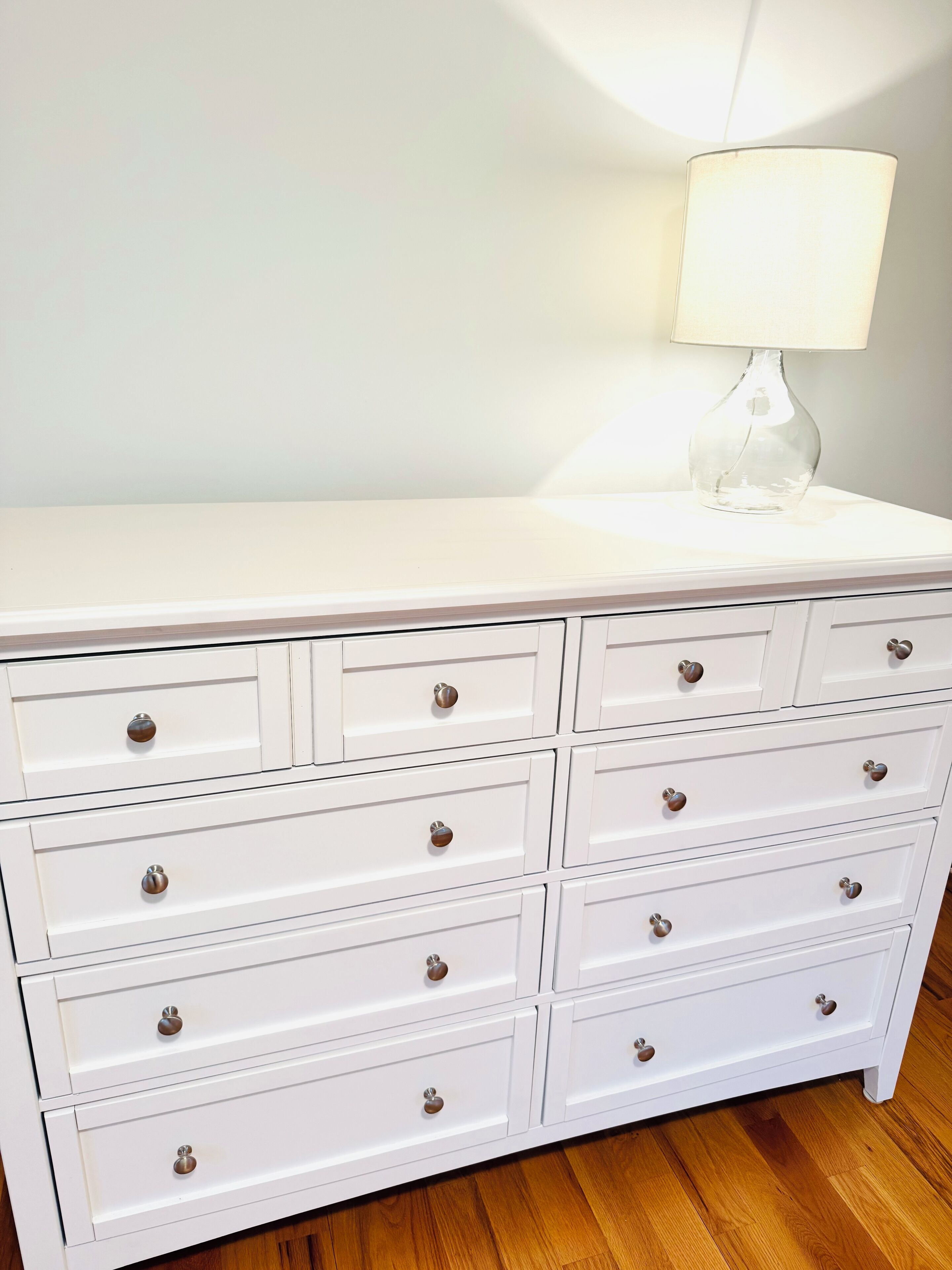 A white dresser with multiple drawers and knobs, a clear glass lamp on top, and a wooden floor.