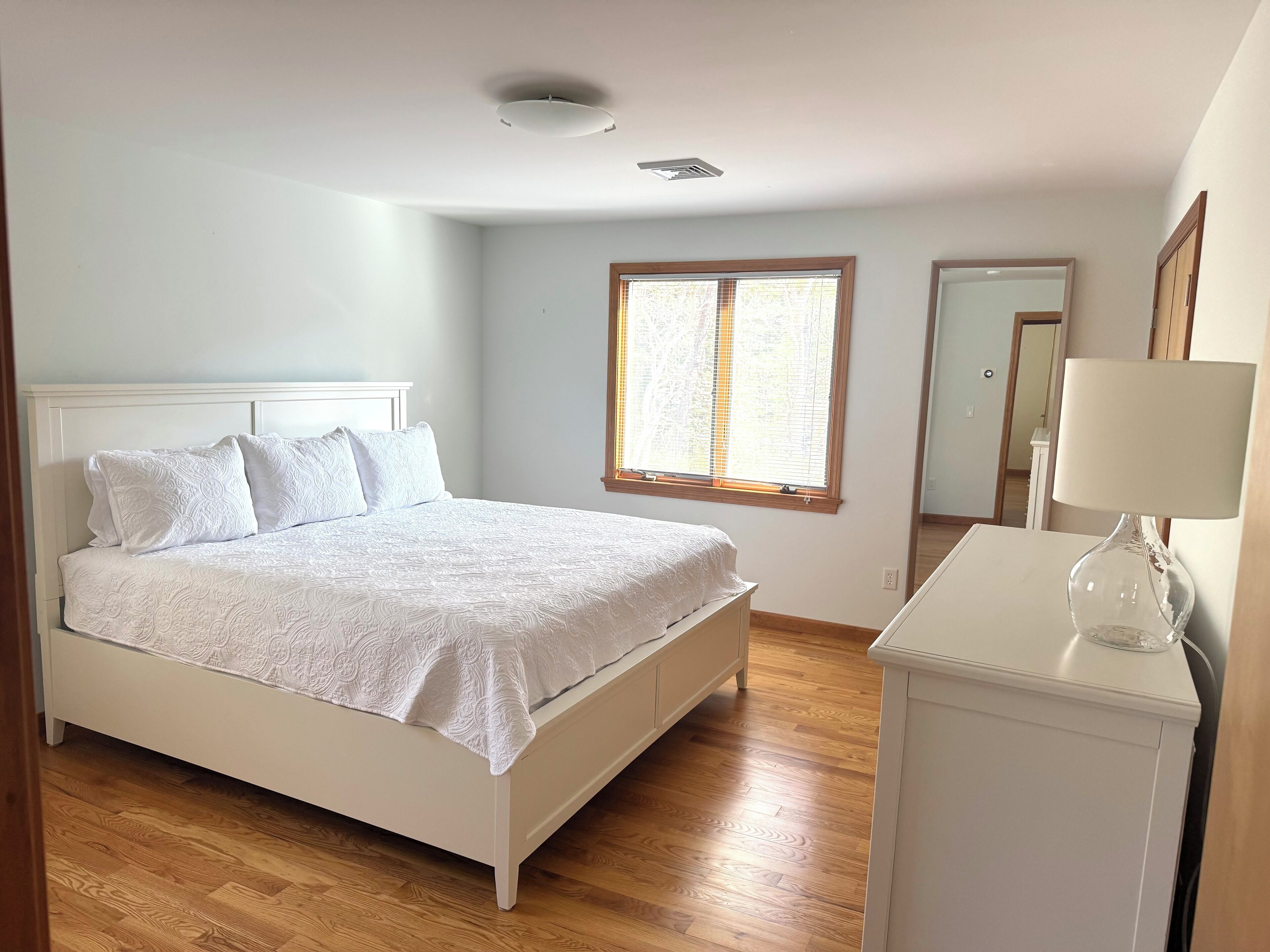 A bedroom with a bed, a bedside table, a window with blinds, a ceiling light, and a mirror.