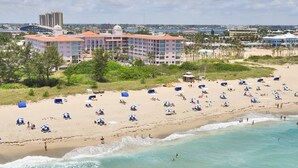 A beach with numerous umbrellas and people. A large hotel with a red roof is visible in the background.