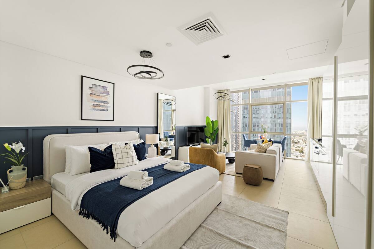 A modern hotel room with a large bed, a desk, a chair, a sofa, and a balcony with a city view.