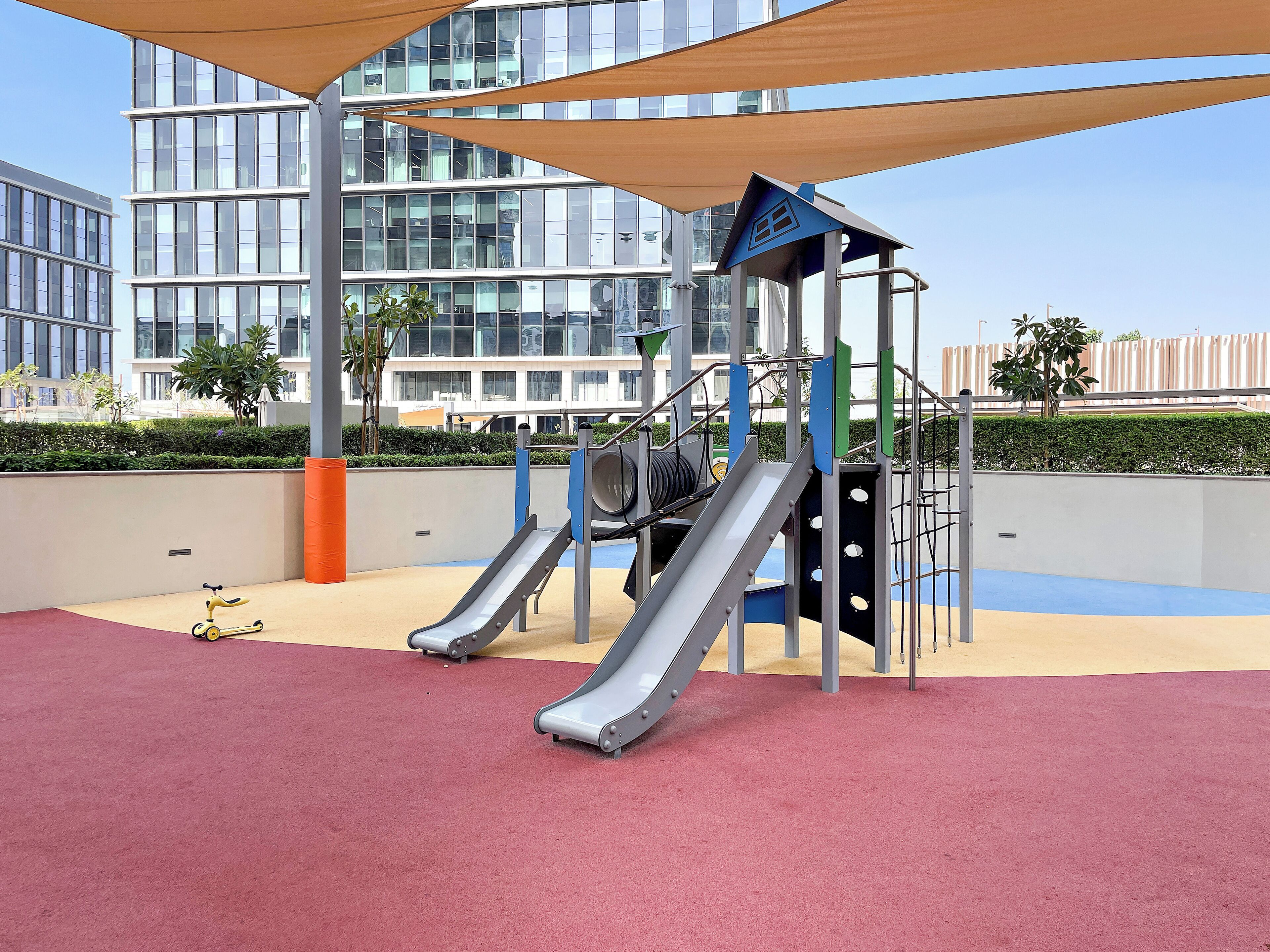 Ein Spielplatz mit zwei Rutschen und einer Kletterkonstruktion vor einem modernen Gebäude mit großen Fenstern.