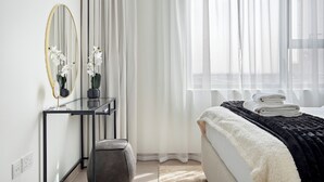 Une chambre à coucher moderne avec un lit, une table de chevet dotée d’un miroir et une fenêtre ornée de rideaux légers.