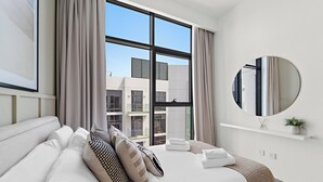 A modern hotel room with a large bed, a round mirror, and a view of the cityscape through large windows.