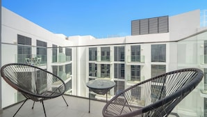 A modern balcony with two wicker chairs and a small table, surrounded by glass railings and white walls.