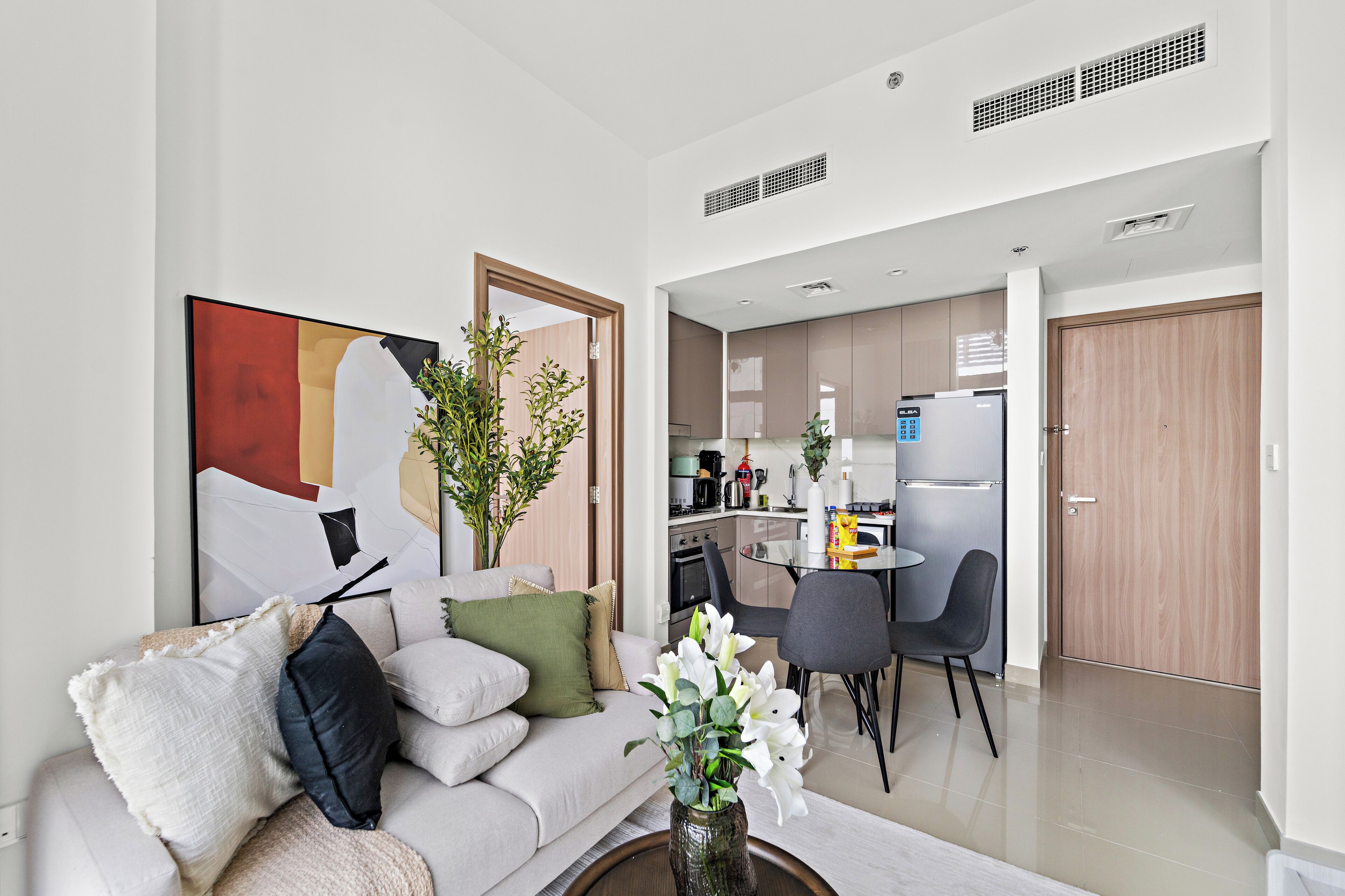 A modern living room with a sofa, a coffee table, and a painting. There is a dining area with a table and chairs. The kitchen is visible in the background with cabinets and appliances.