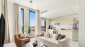 Een moderne woonkamer met een bank, fauteuil en salontafel, een keuken met lichte houten kastjes, en een stadszicht door grote ramen.