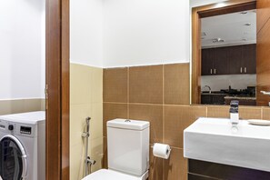 A bathroom with a washing machine, toilet, sink, and a mirror reflecting a kitchen area. - Sillkhaus Yasmine Tower (Abu Dhabi)