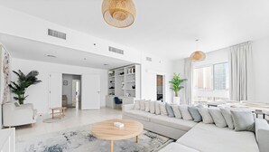 En rummelig stue med en stor sofa, et rundt sofabord og et spiseområde med bord og stole. Der er et køkken i baggrunden, og rummet er holdt i en moderne, minimalistisk stil.