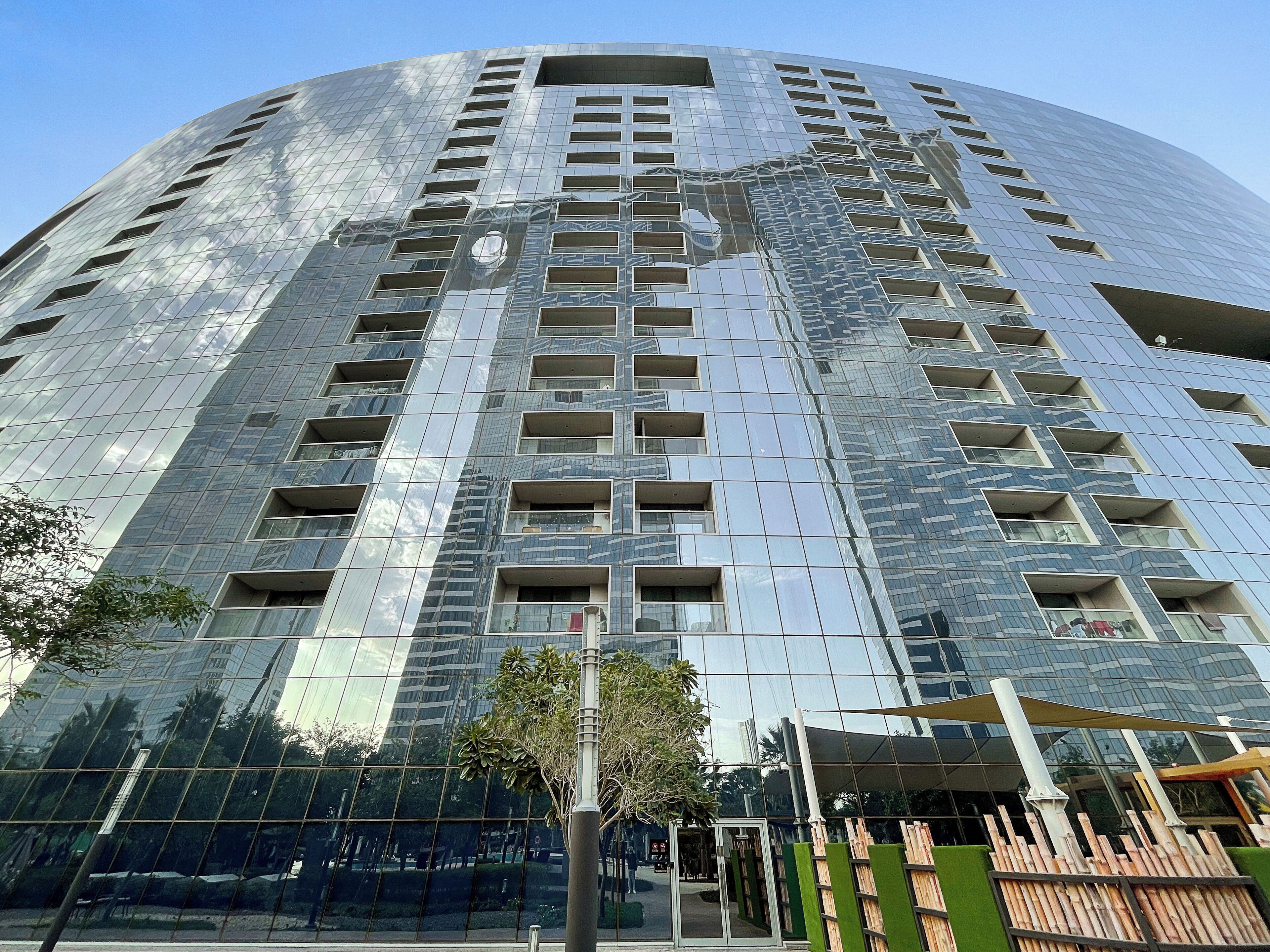 Een modern hoog gebouw met een glazen gevel die reflecteert, meerdere balkons en een centrale ingang.