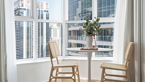 Une pièce avec une table ronde et deux chaises en osier, un vase avec une plante, et une vue sur de hauts immeubles à travers de grandes fenêtres.