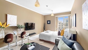 A modern hotel room with a large bed, a sofa, a dining area with chairs, a TV, and a city view. - Silkhaus Goldcrest Executive (Dubai)