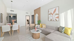 A modern living room with a grey sofa, a round coffee table, and a dining area with a white table and chairs. - Silkhaus Goldcrest Executive (Dubai)