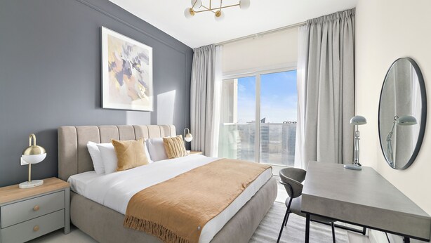 A modern hotel room with a large bed, a desk, a chair, a bedside table with lamps, a large window with curtains, and a ceiling light fixture. - Silkhaus Goldcrest Executive (Dubai)