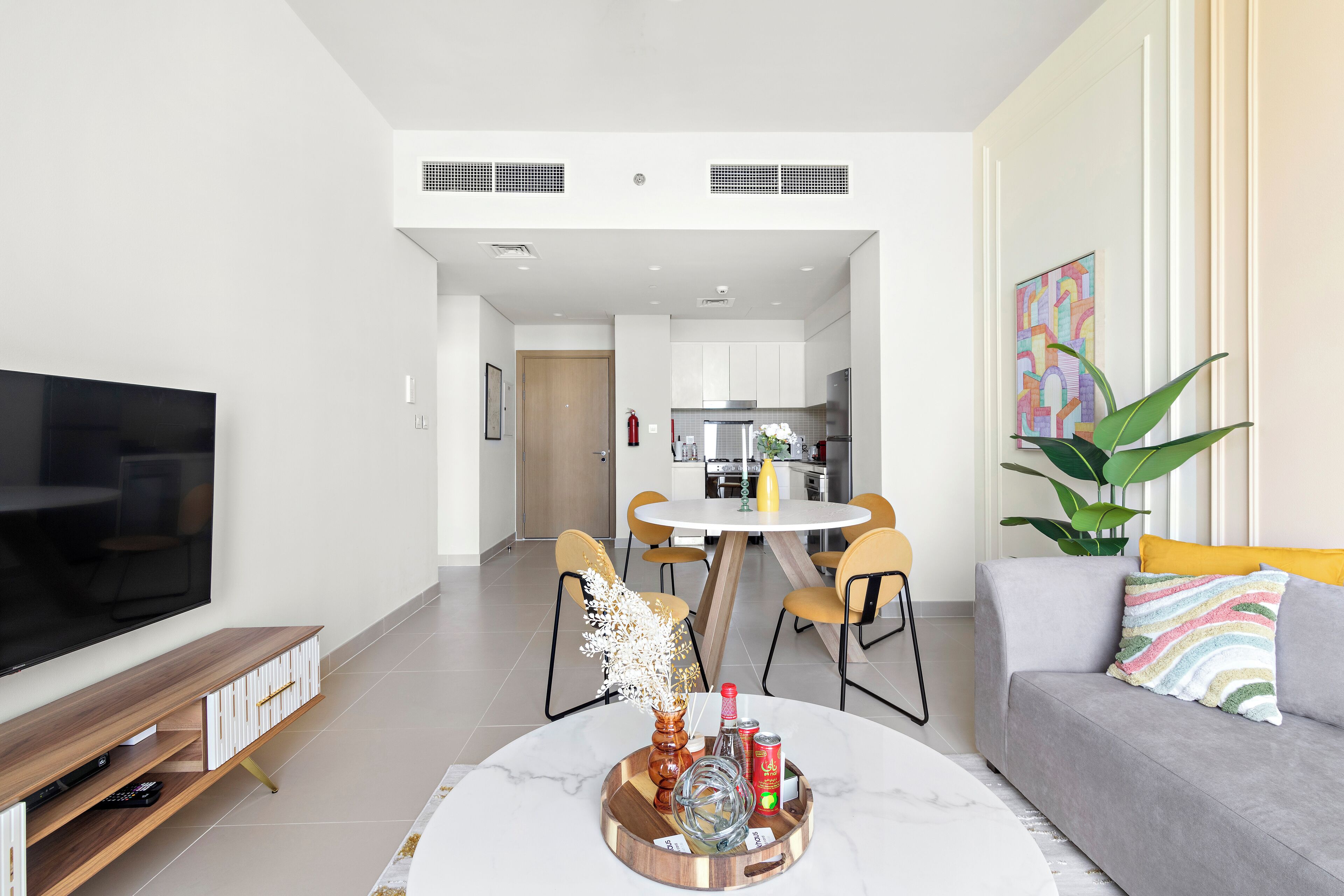 A modern living room with a flat-screen TV, a coffee table with decorative items, a grey sofa, and a dining area with yellow chairs.