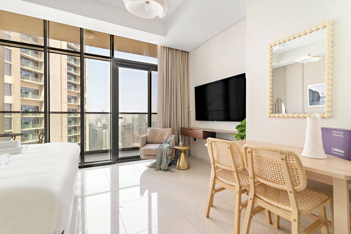 A modern hotel room with a large window, a flat-screen TV, a bed, a desk with a chair, and a mirror.