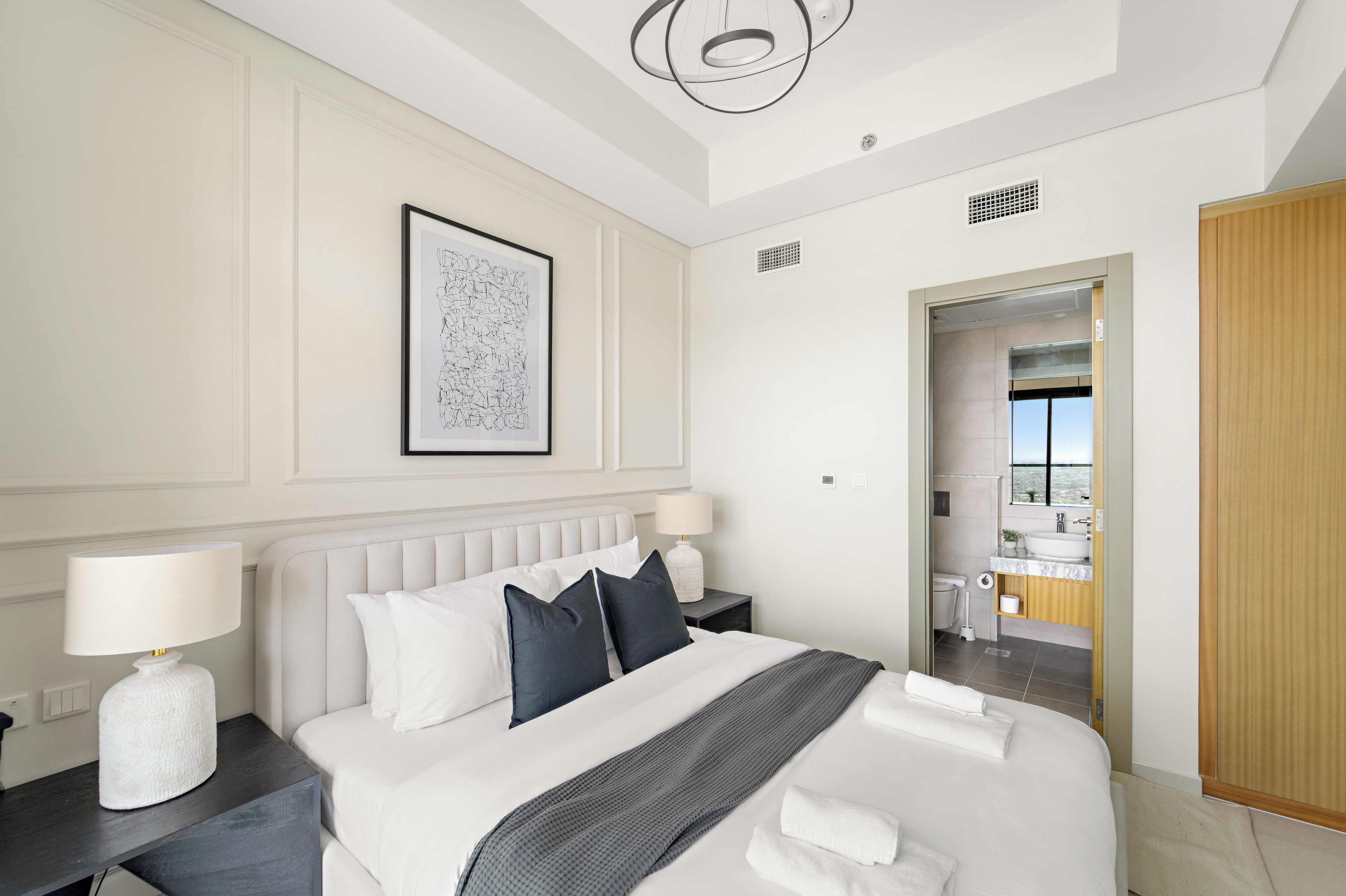 A modern hotel room with a large bed, white bedding, a framed artwork on the wall, and a view of the bathroom through an open door.