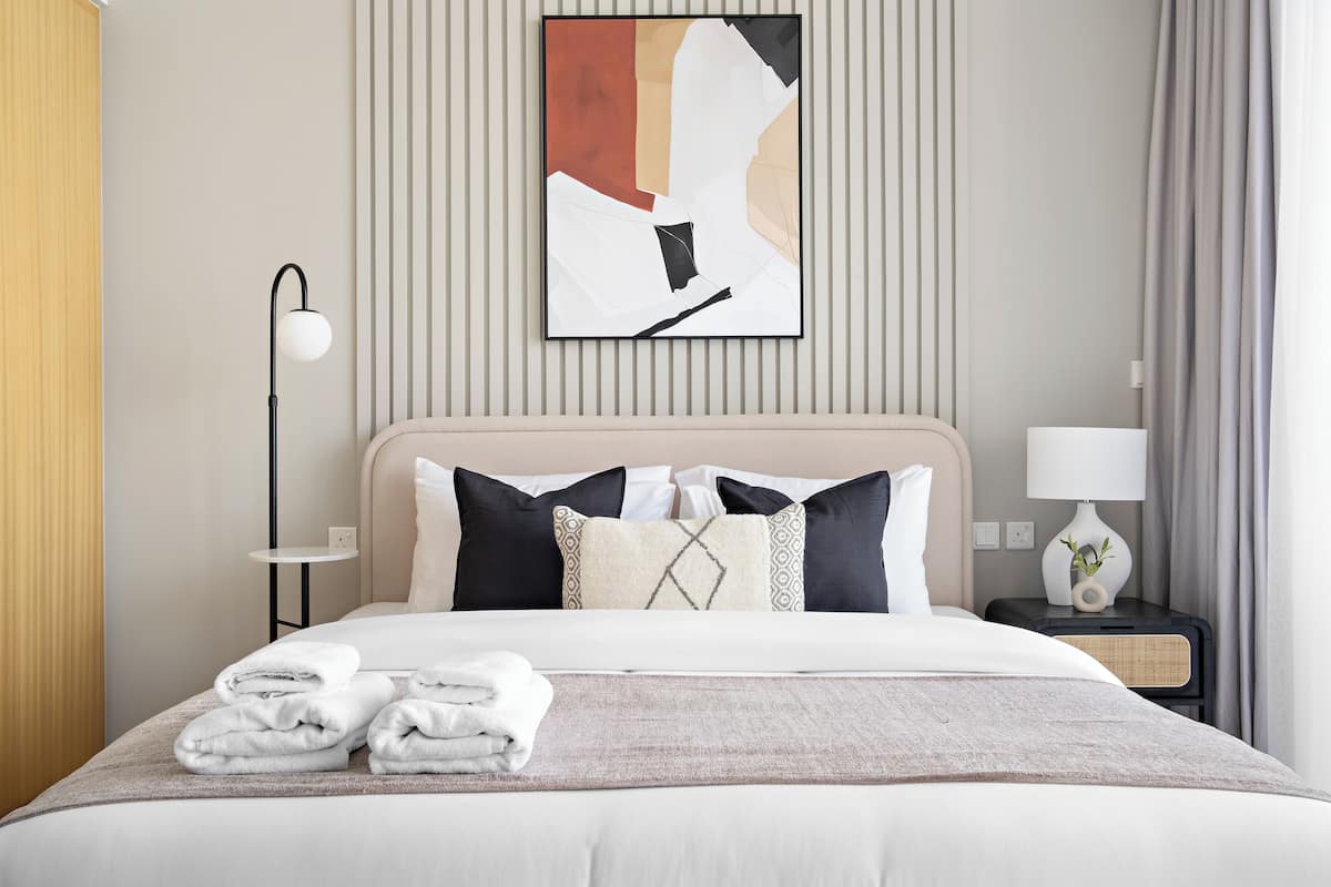 A neatly made bed with a white comforter and several pillows, a bedside table with a lamp and a small plant, and a wall-mounted abstract painting.
