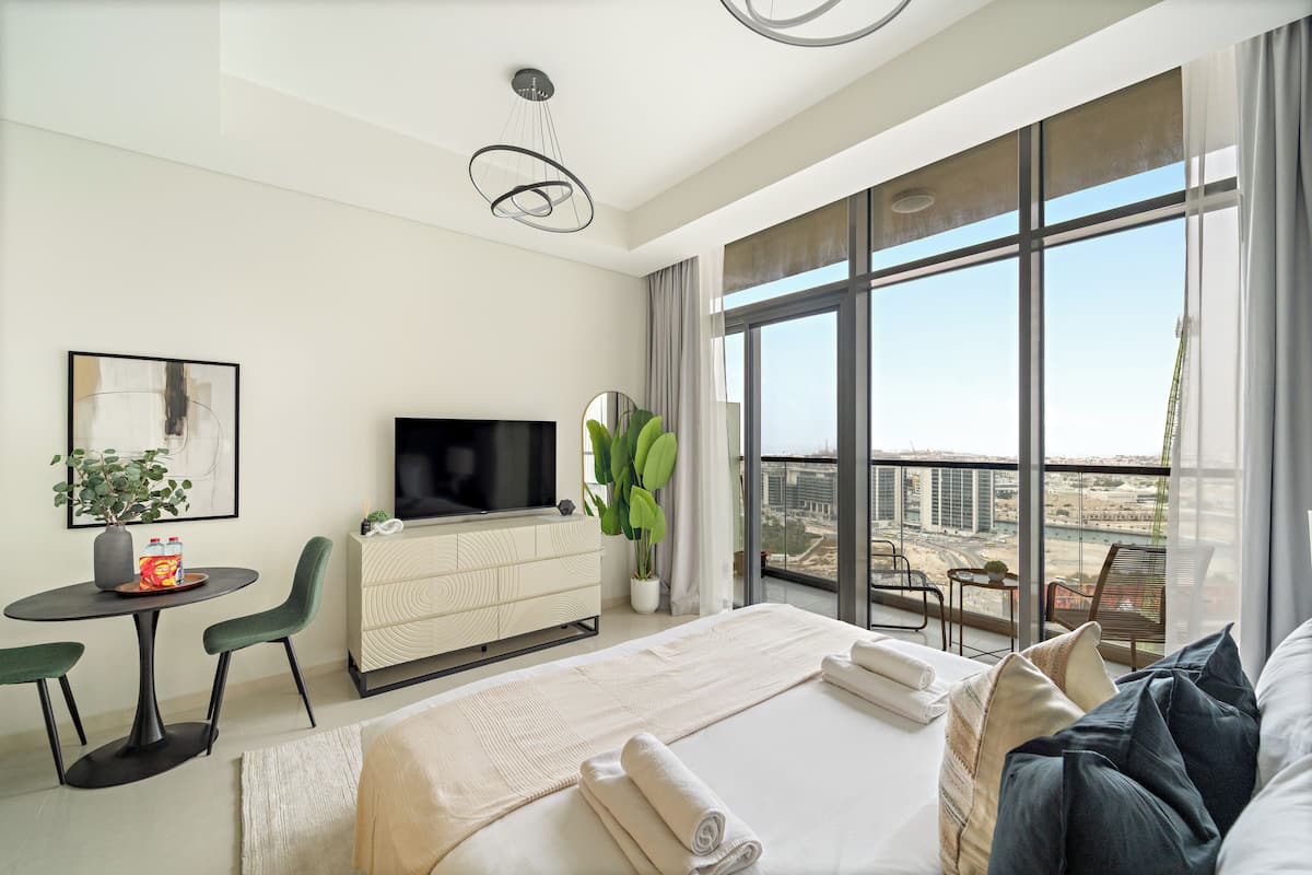 A modern hotel room with a large bed, a TV, a dining area with a table and chairs, and a balcony with a view of the city.