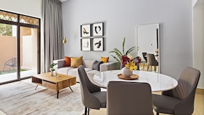 Un moderno salón con sofá, mesa de centro y zona de comedor con sillas. Hay un ventanal con cortinas, una lámpara de araña y cuadros en la pared.