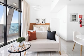 A modern hotel room with a large window, a bed with white and black pillows, a small round table with a plant, and a white chair.