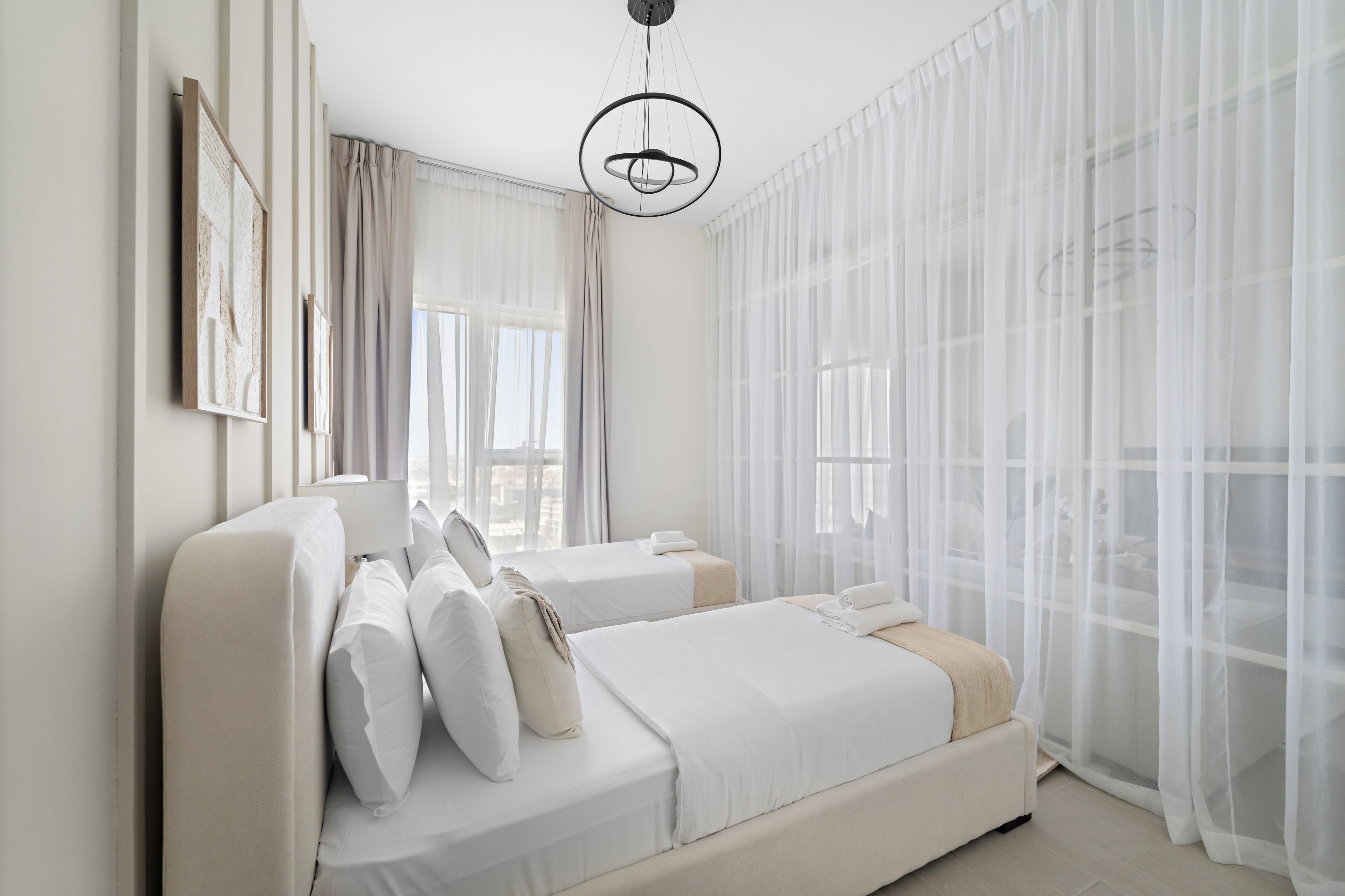 A hotel room with two beds, a large window with sheer curtains, and a modern chandelier.