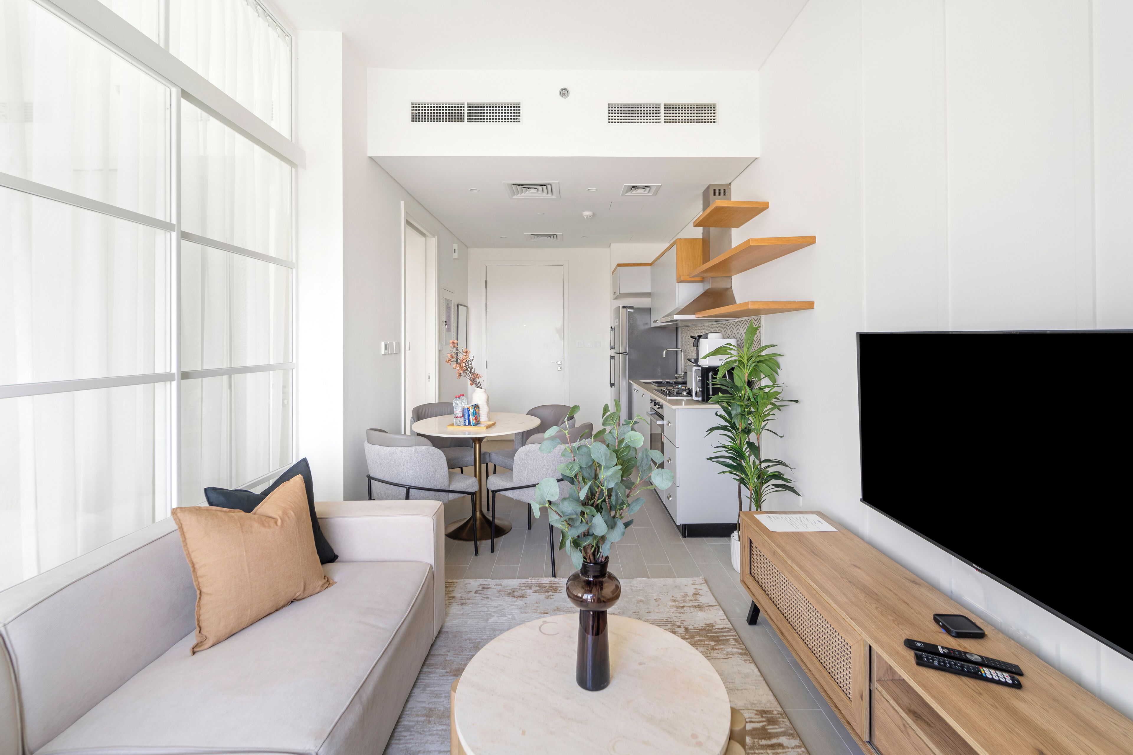A modern living room with a sofa, coffee table, and TV. There is a dining area with chairs and a table, and a kitchen area with a stove and cabinets.