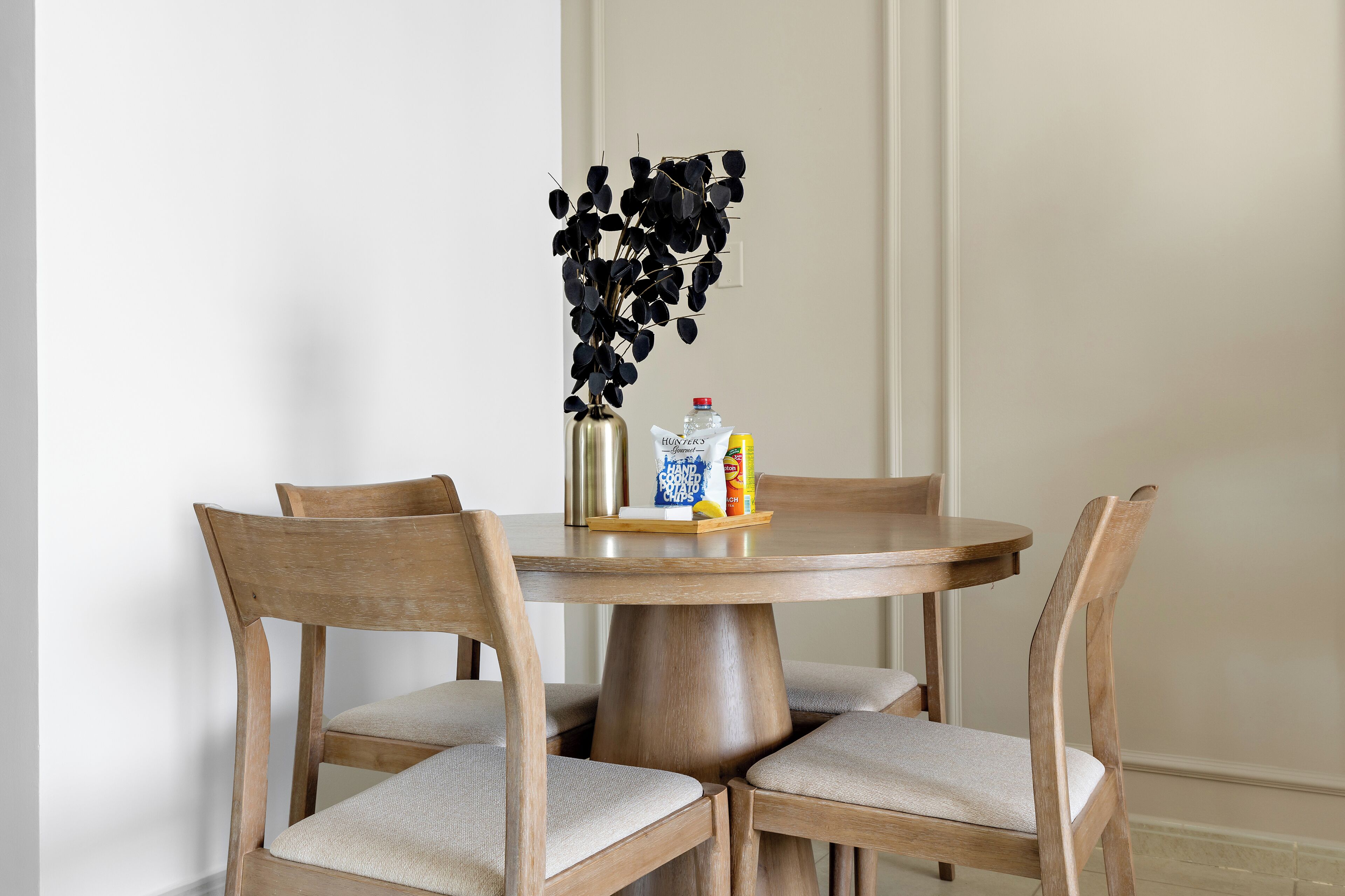 A round wooden dining table with four chairs, a vase with black leaves, a bottle of hand soap, and a box of snacks.