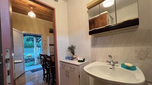 Ein Badezimmer mit Waschbecken, Spiegel und Schränken. Eine Tür führt zu einem Essbereich mit Stühlen und einem Tisch.