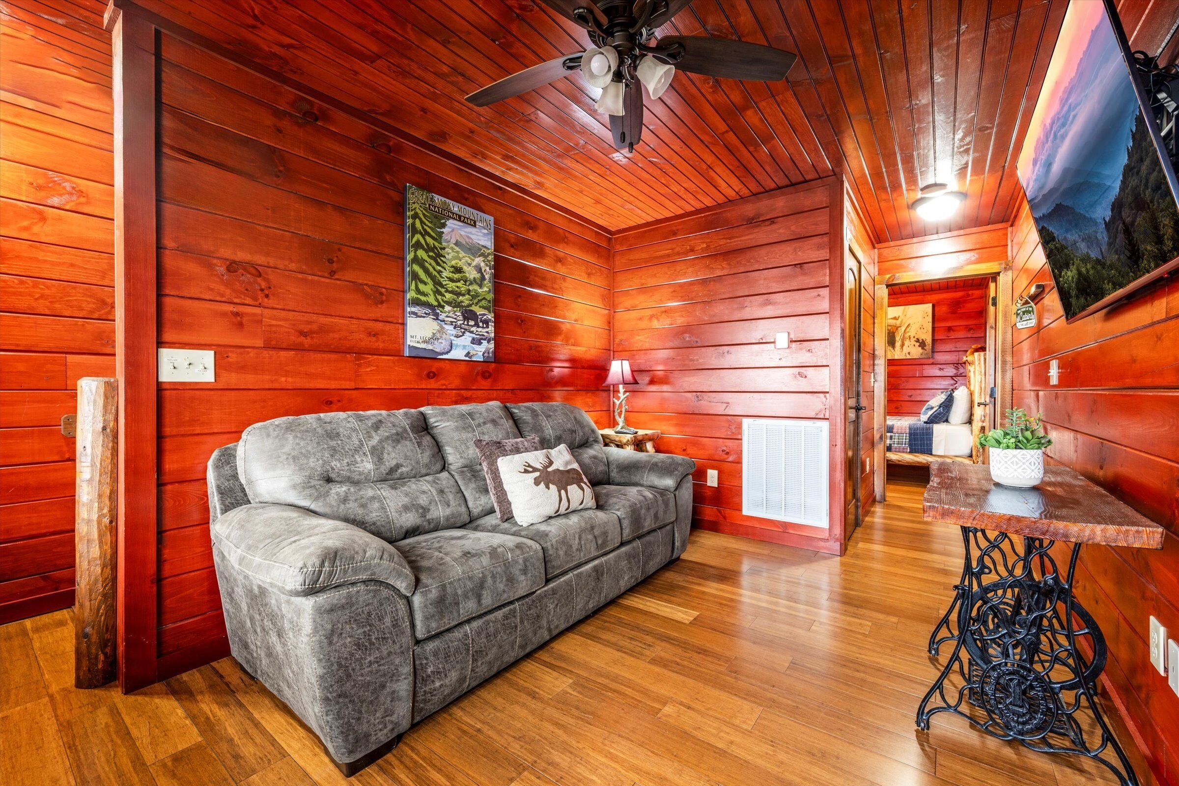 Une pièce chaleureuse aux murs en bois, avec un canapé gris orné d’un coussin à l’effigie d’un élan, un plancher en bois, un ventilateur de plafond et un tableau représentant un paysage de montagne.