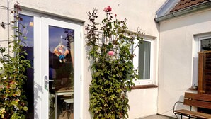 Una casa con fachada blanca, puerta corrediza de vidrio, un pequeño jardín con un banco y un rosal.