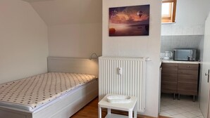 Een kleine, compacte kamer met een bed, een tafeltje, een radiator, een magnetron en een kitchenette.