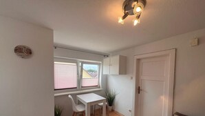 Een lichte, kleine kamer met een witte tafel en stoelen, een potplant, een witte deur en een raam met jaloezieën.
