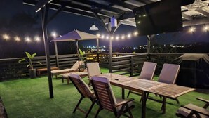 An outdoor patio with a wooden table and chairs, a pergola with string lights, and a view of the city at night. - Villa Boho Motorhome (Cabo Rojo)