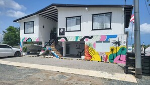 A two-story building with a white exterior and black window frames. There is a staircase leading to the entrance, and a colorful mural on the side of the building. - Villa Boho Motorhome (Cabo Rojo)