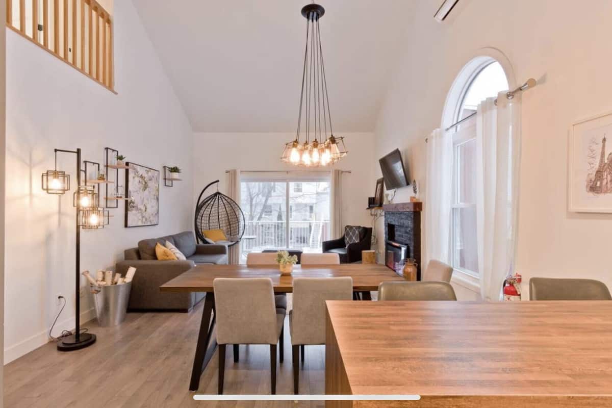 Un salon moderne avec un coin repas, comprenant une table à manger en bois, des canapés gris, un luminaire suspendu et une cheminée.