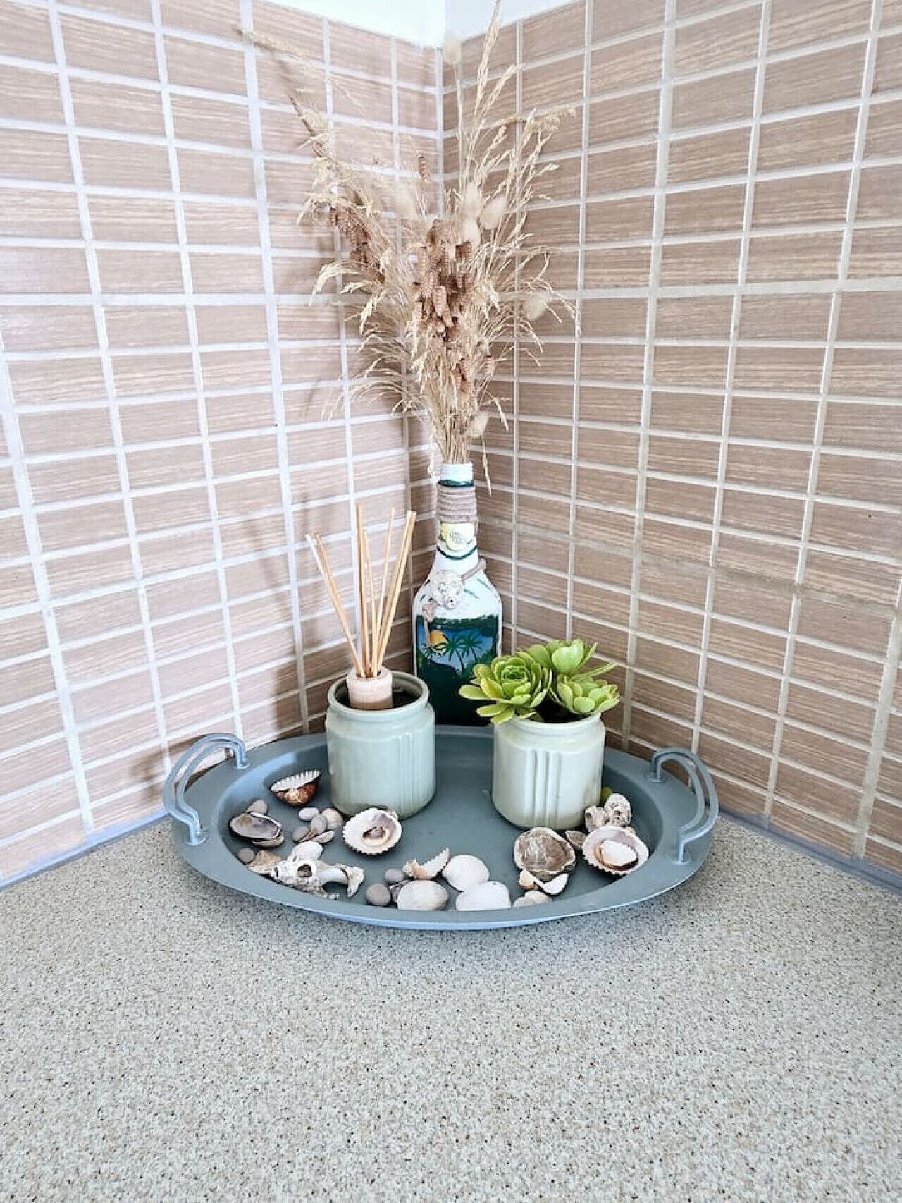 Un coin avec un plateau gris contenant des coquillages, une plante succulente verte et des plumes de jarre séchées dans une bouteille.
