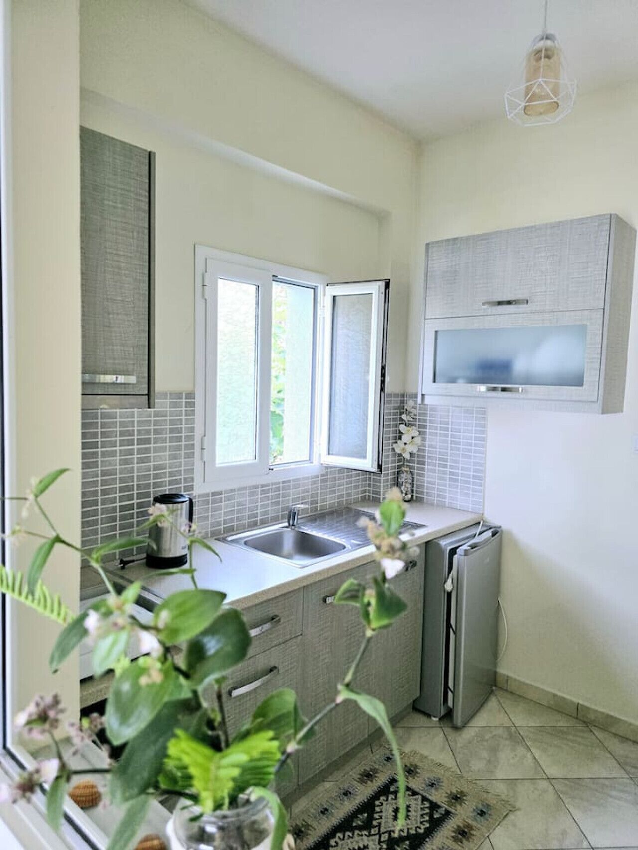 A compact kitchen with a built-in microwave, a sink, and a refrigerator. There are two windows providing natural light, and a decorative plant is visible in the foreground.
