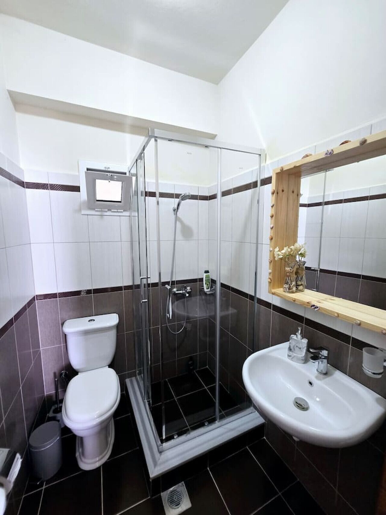 Un baño con ducha de vidrio, inodoro, lavamanos y espejo con repisa de madera.