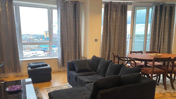 A living room with a sofa, ottoman, and a dining table with chairs. - Cosy 2 Bedroom Royal Docks Apartment (London)