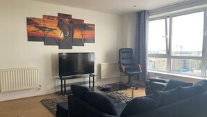 A living room with a large flat-screen TV, a black leather chair, a dark-colored sofa, and a city view through large windows. - Cosy 2 Bedroom Royal Docks Apartment (London)