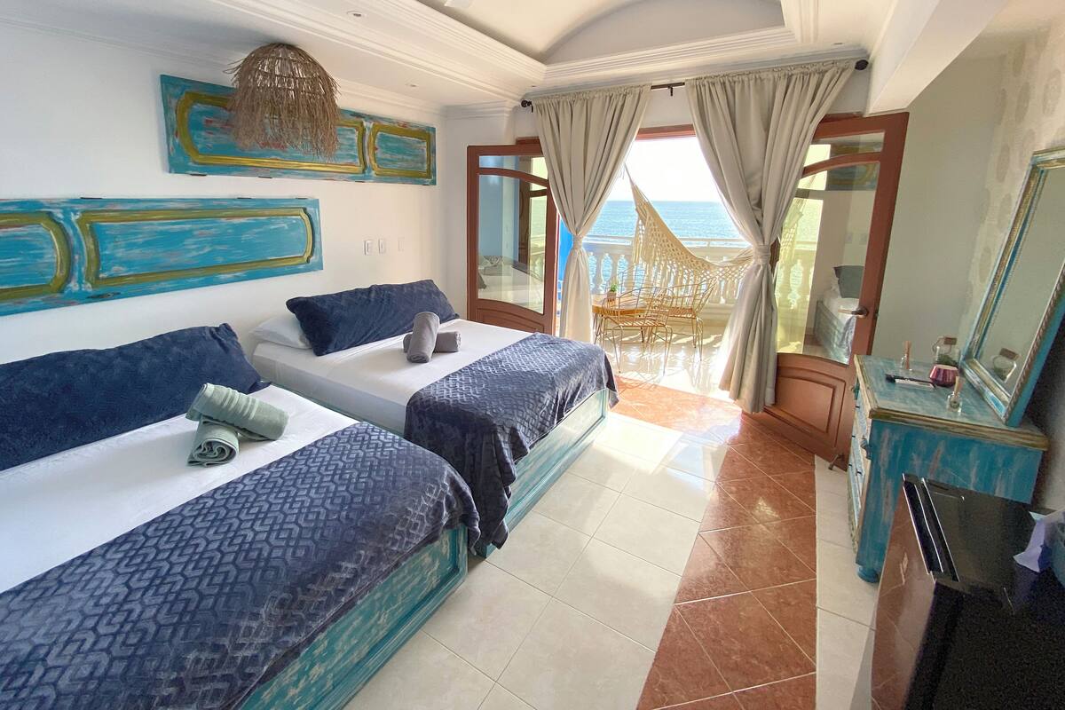 A hotel room with two beds, a large window with a view of the sea, a balcony with a wicker chair, and a vanity with a mirror.
