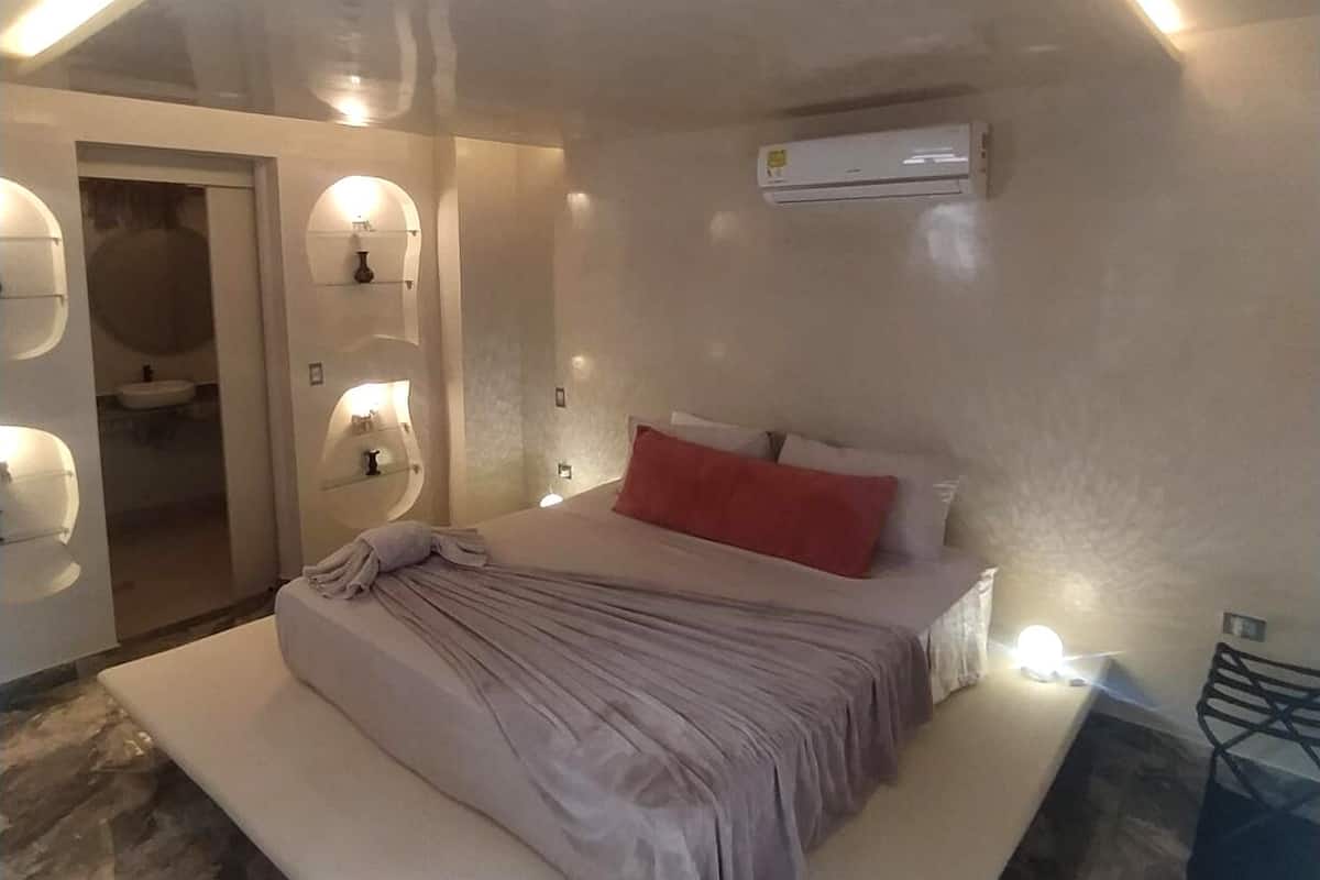 A modern bedroom with a large bed, a built-in bench, a bedside table, an air conditioning unit, and a bathroom visible through an open door.