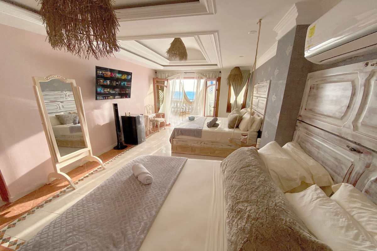 A spacious bedroom with a large bed, a sitting area, a TV, and a view of the sea.