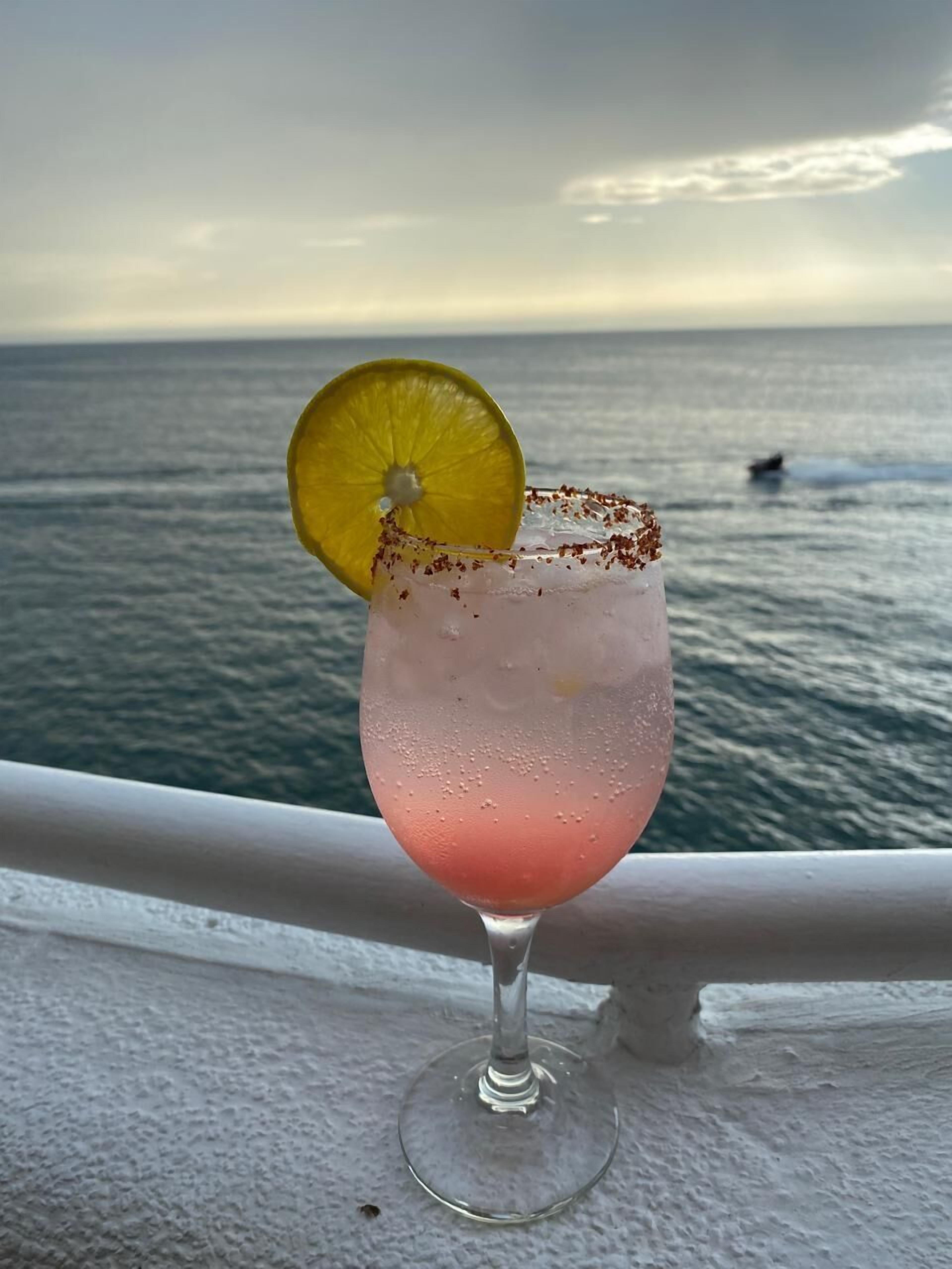 A pink cocktail with a lime garnish and salt rim, served in a glass on a balcony overlooking the sea.