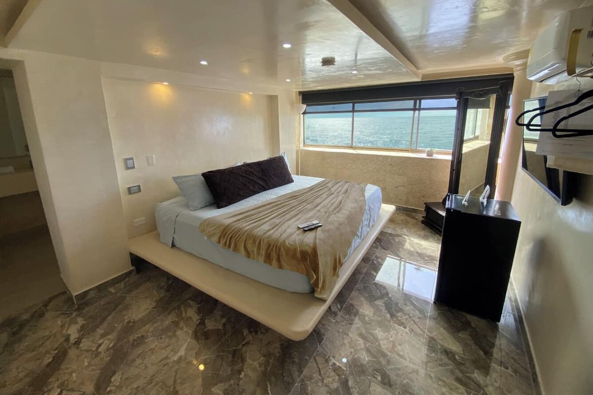 A bedroom with a bed, a bench, a desk, and a large window with a view of the sea.