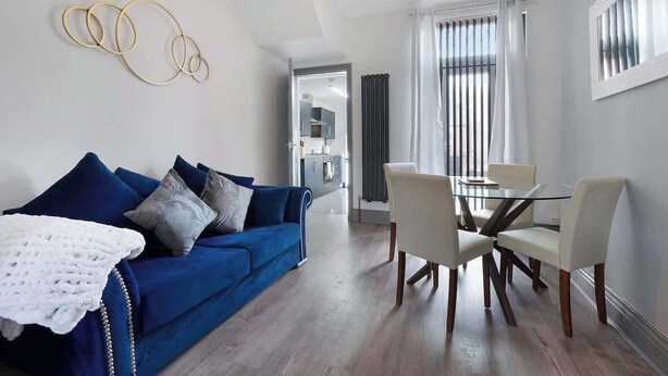 A modern living room with a blue sofa, a glass dining table with chairs, and a kitchen in the background. - Comfortable 2BR Home 10 Min to Durham Cathedral (Ushaw Moor)